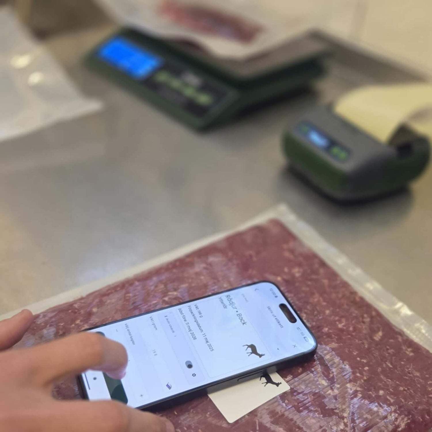 En hand använder en smartphone för att hantera viltkött med Tendy etikettskrivare och vågpaket i bakgrunden.